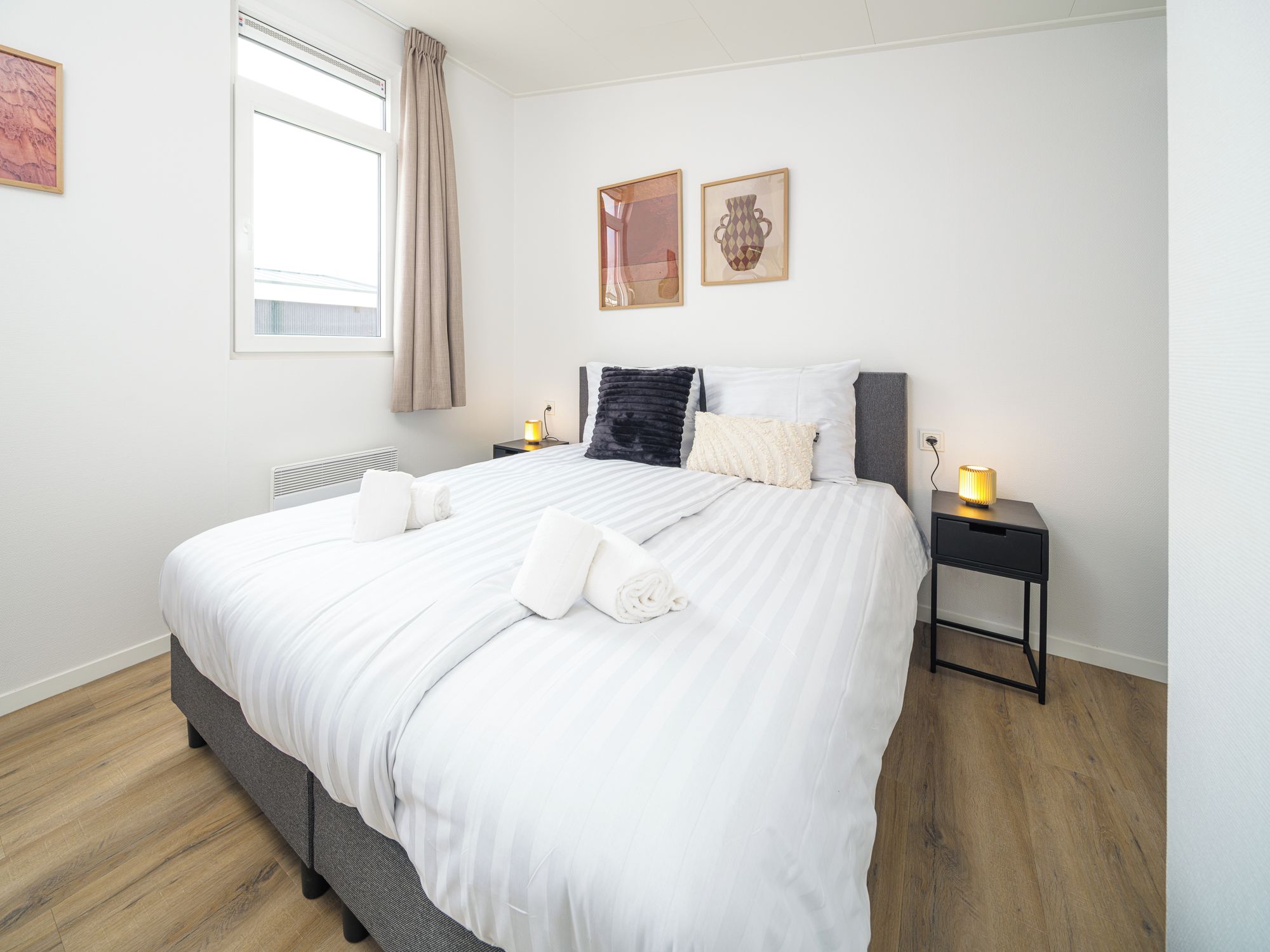 Un lit bien fait, avec du linge de lit blanc et une serviette pliée, une table de chevet avec une lampe et une fenêtre avec des rideaux.