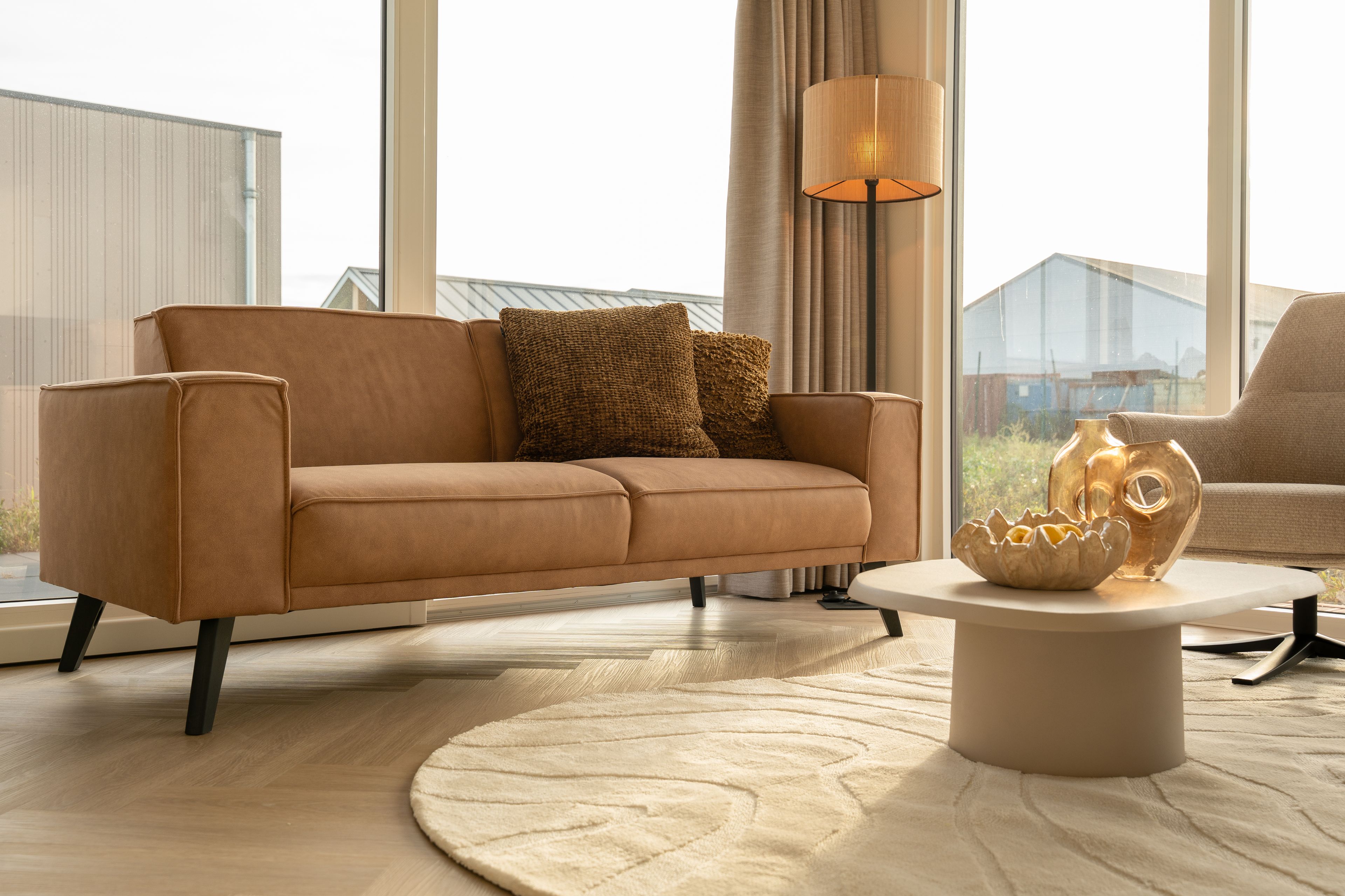 Een moderne woonkamer met een bruine bank, een ronde salontafel en een lamp.