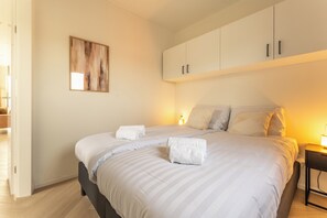 Una cama bien hecha con sábanas blancas y dos toallas, una mesita de noche con lámpara, y un cuadro en la pared.