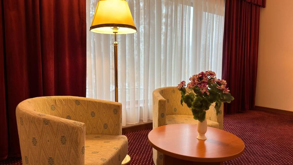 A hotel room with two armchairs, a small round table, a vase with flowers, a floor lamp, and a window with curtains.