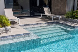 En swimmingpool med fliserede kanter, liggestole og en stenvæg.