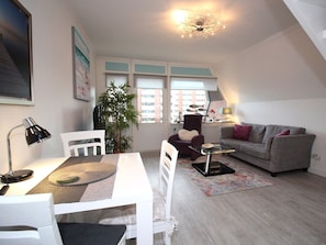 A modern living room with a dining area, a sofa, a chair, a lamp, and a plant. - Atrium Holiday Apartment 33 (Sylt)