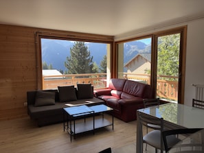 A room with a wooden floor, a red leather sofa, a glass coffee table, and a dining table with chairs. Large windows offer a view of mountains and trees. - Apartment with balcony and ski slopes view (Les Deux Alpes)