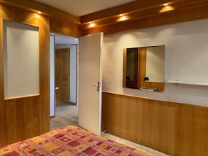 Une chambre avec des lambris en bois, un lit recouvert d’une couvre-lit à motifs, une table de chevet, un miroir et une porte donnant sur une autre pièce.