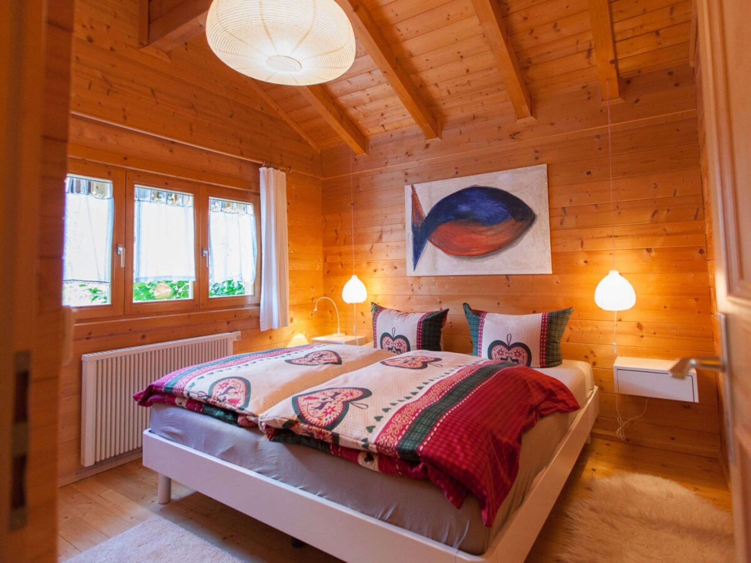 A wooden-paneled room with a bed, two bedside lamps, a radiator, and a window with curtains.