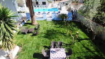 Zona de piscina con sillones de descanso y una mesa, rodeada de césped cuidado y palmeras.