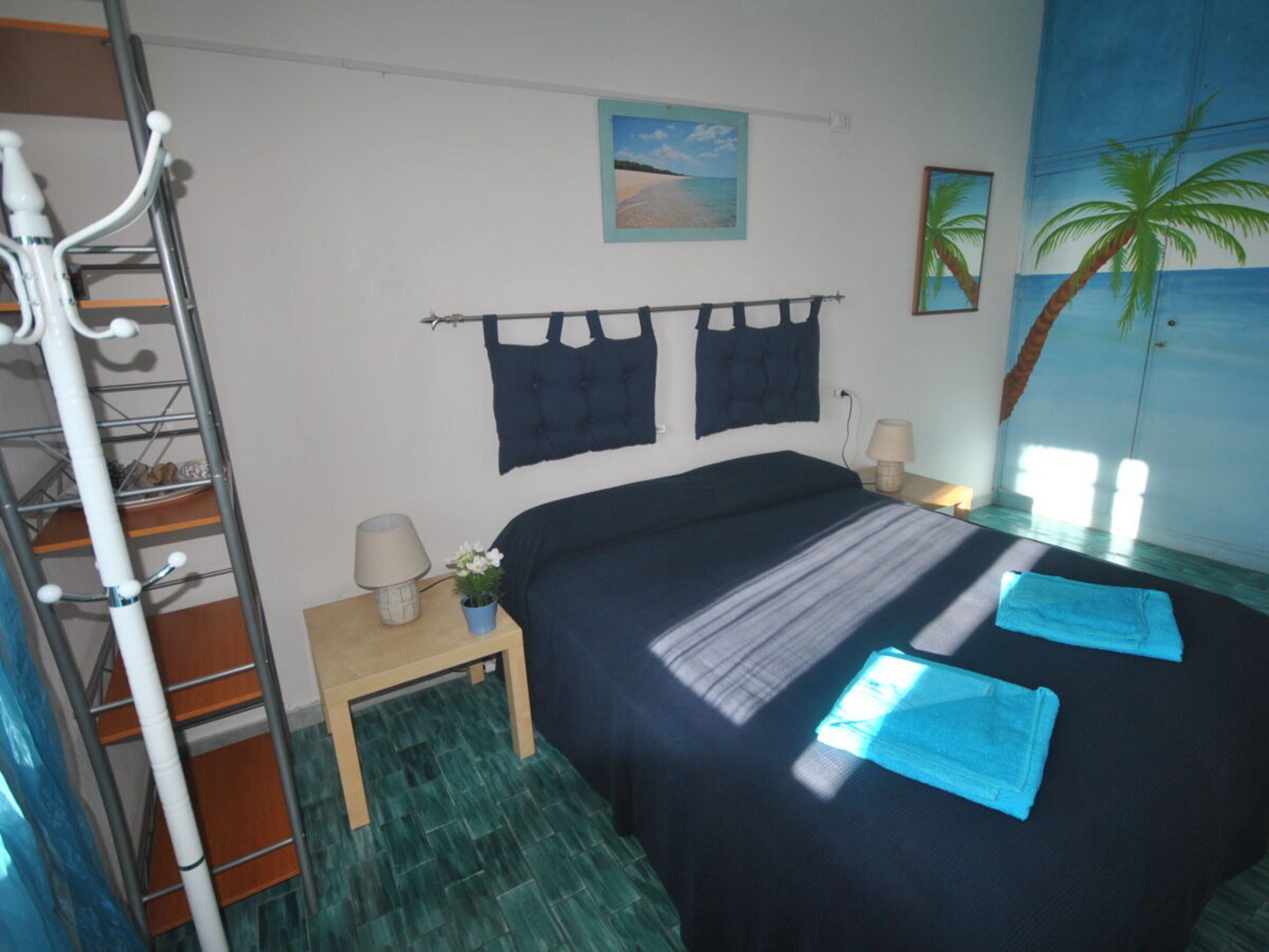 Un dormitorio con una cama, dos lámparas de pared, una mesita de noche, una pequeña mesa, una planta en maceta y un cuadro con una escena de playa.