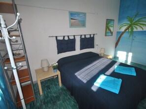 A bedroom with a bed, two wall-mounted lights, a nightstand, a small table, a potted plant, and a painting of a beach scene. - Holiday Apartment With 1 Bedroom (Orosei)