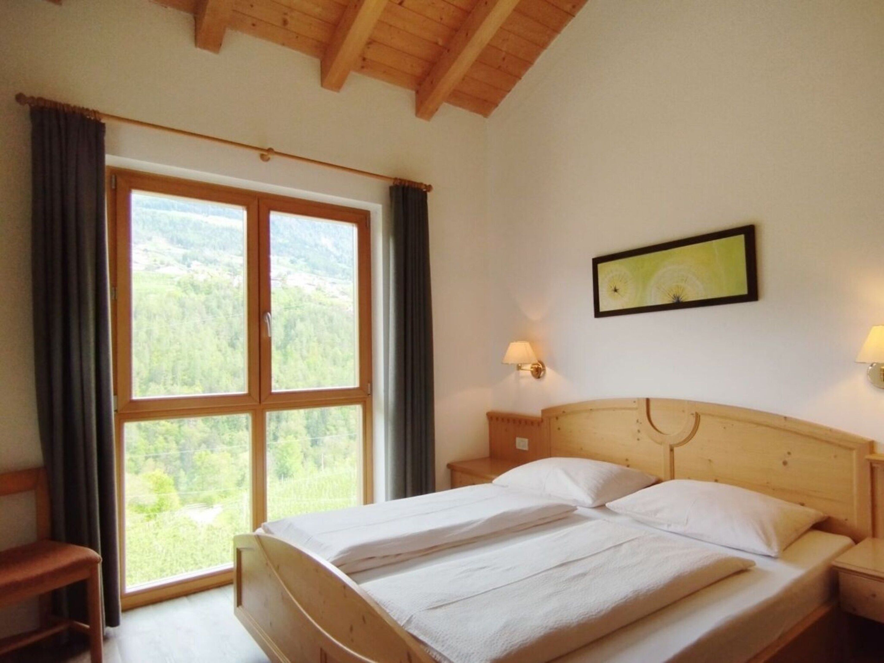Una camera da letto con soffitto in legno, un'ampia finestra con tende e un letto con lenzuola bianche, oltre a un lampadario a parete.