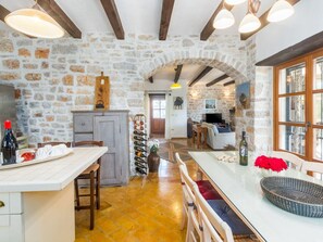 Eine rustikale Küche mit Steinwänden, Holzbalken, einem Essbereich mit langem Tisch und einer gemütlichen Wohnstube mit Kamin.
