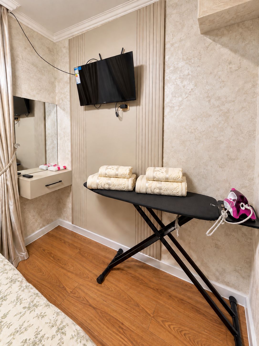 Una habitación de hotel con piso de madera, una cama con ropa de cama estampada, una tabla de planchar negra con toallas, un televisor montado en la pared y un espejo.