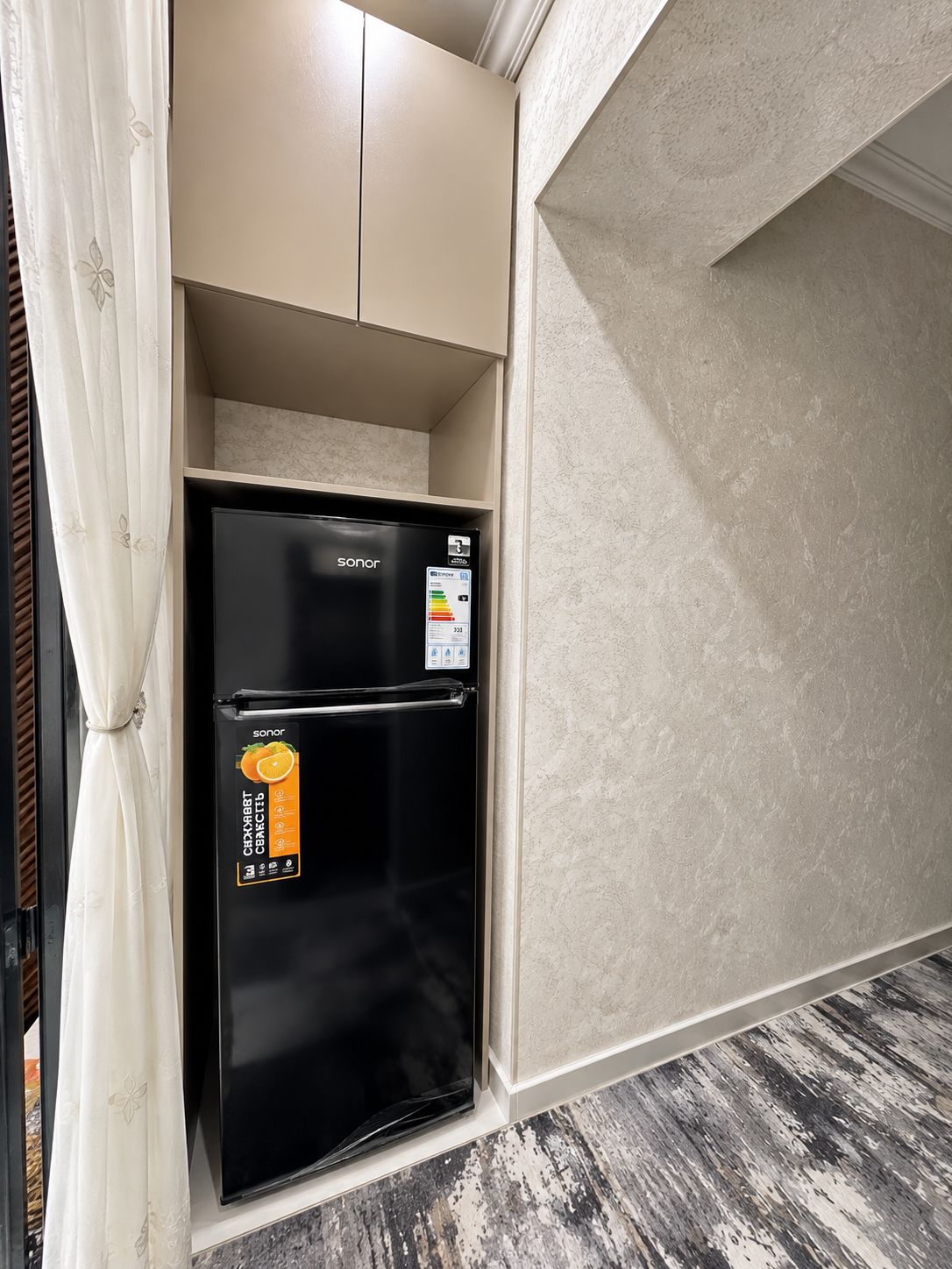 Ein schwarzer Kühlschrank mit einem Aufkleber, ein beiges Schränkchen darüber und eine strukturierte Wand.