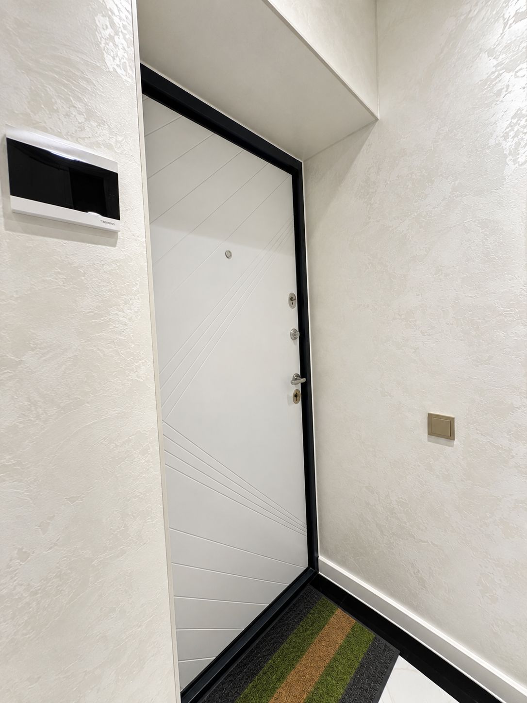Una puerta blanca con marco negro, una rejilla rectangular pequeña y un felpudo con diseño a rayas.