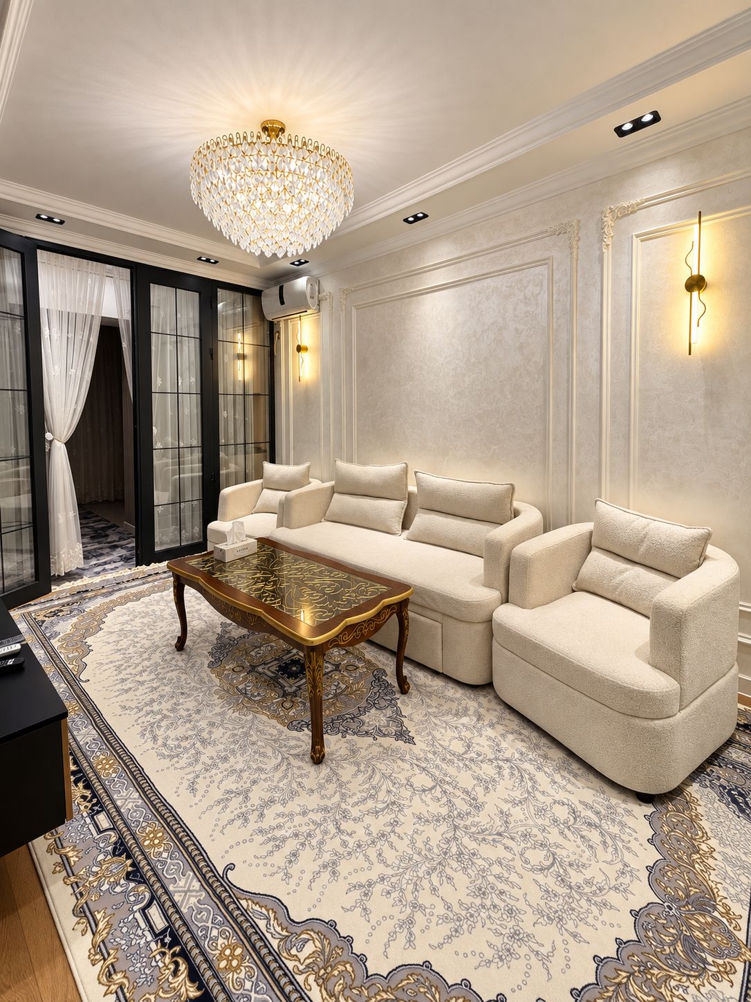 Ein luxuriöses Wohnzimmer mit einem großen Kronleuchter, einem Ecksofa, einem Glastisch und einem gemusterten Teppich.