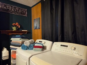 Een wasruimte met gestapelde handdoeken, een wasmachine, een plank met bloemen en een decoratief bord.