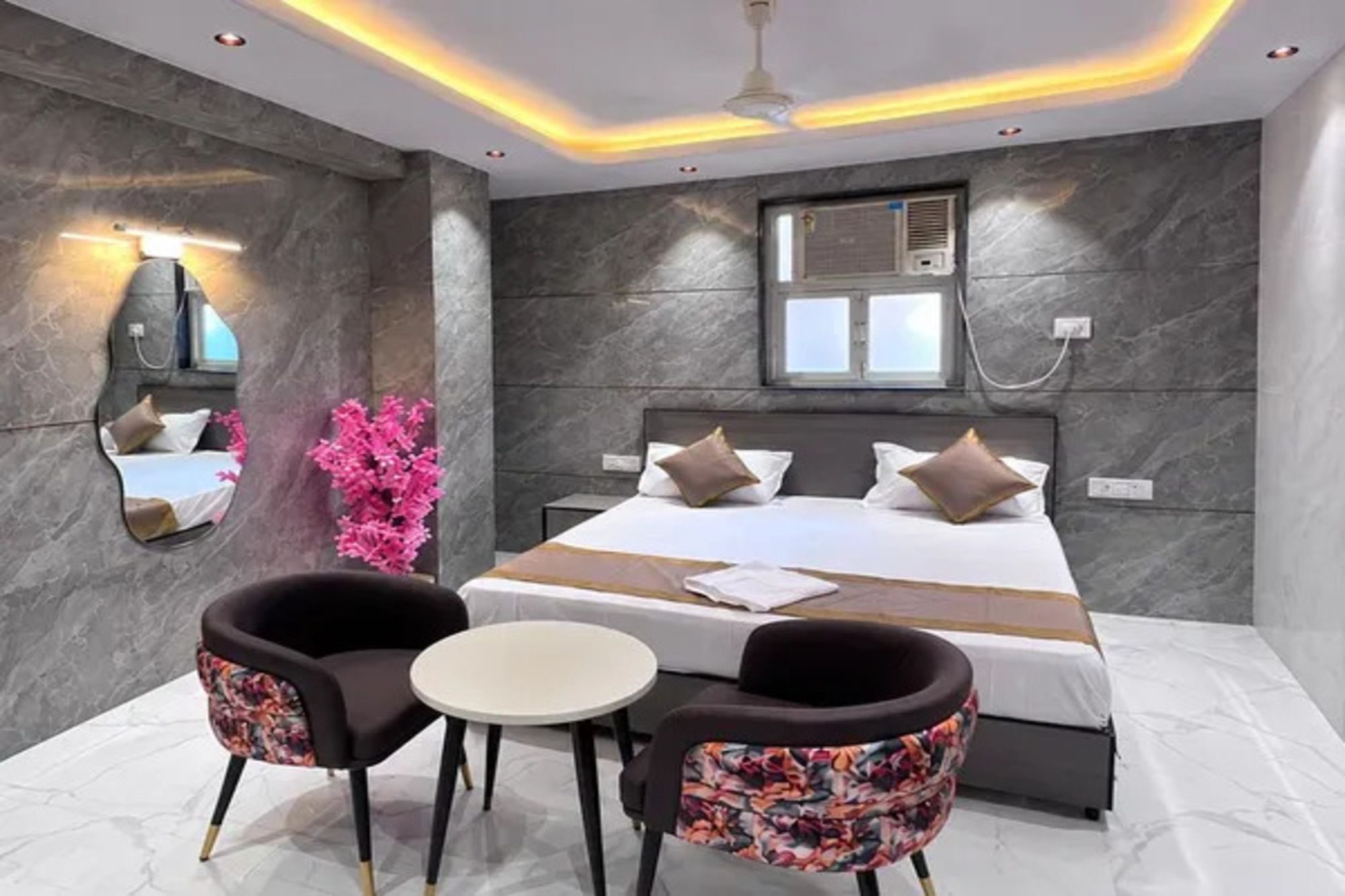 A modern bedroom with a large bed, two chairs, a small table, a mirror, and a window with an air conditioning unit.