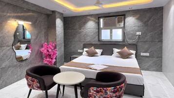A modern bedroom with a large bed, two chairs, a small table, a mirror, and a window with an air conditioning unit.