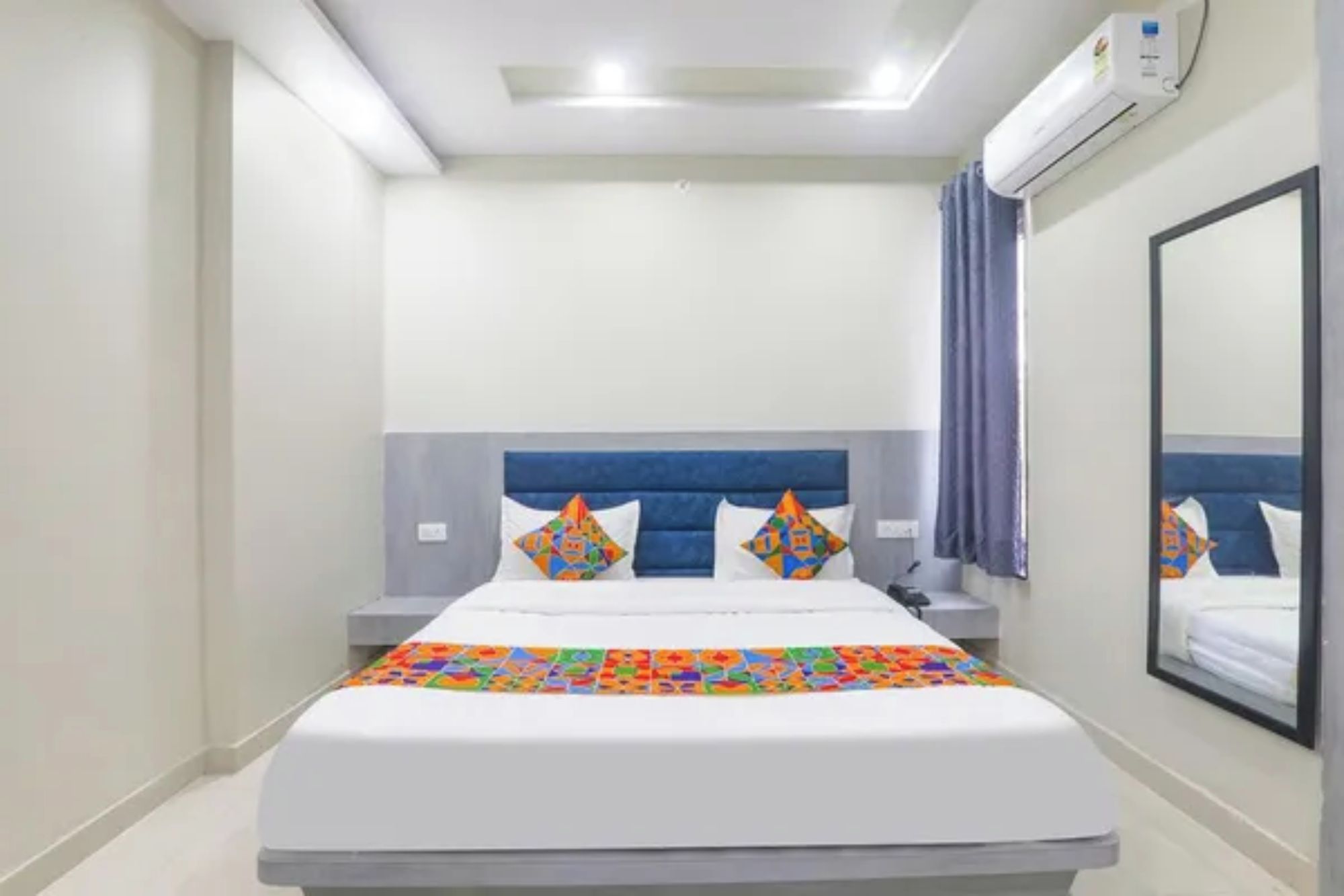 Una habitación de hotel con una cama, una mesita de noche, un equipo de aire acondicionado y un espejo grande en la pared.