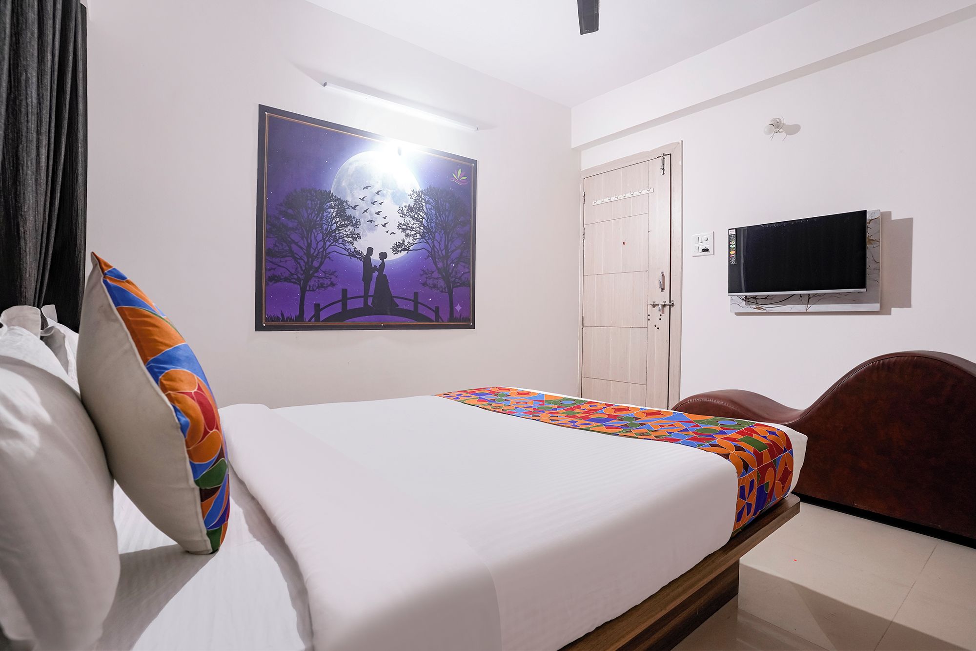 Un dormitorio con cama, televisor, puerta y una pared con un cuadro enmarcado.