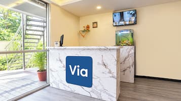 Un mostrador de recepción con diseño de mármol y el logo 'Via', un televisor montado en la pared y una planta en maceta.