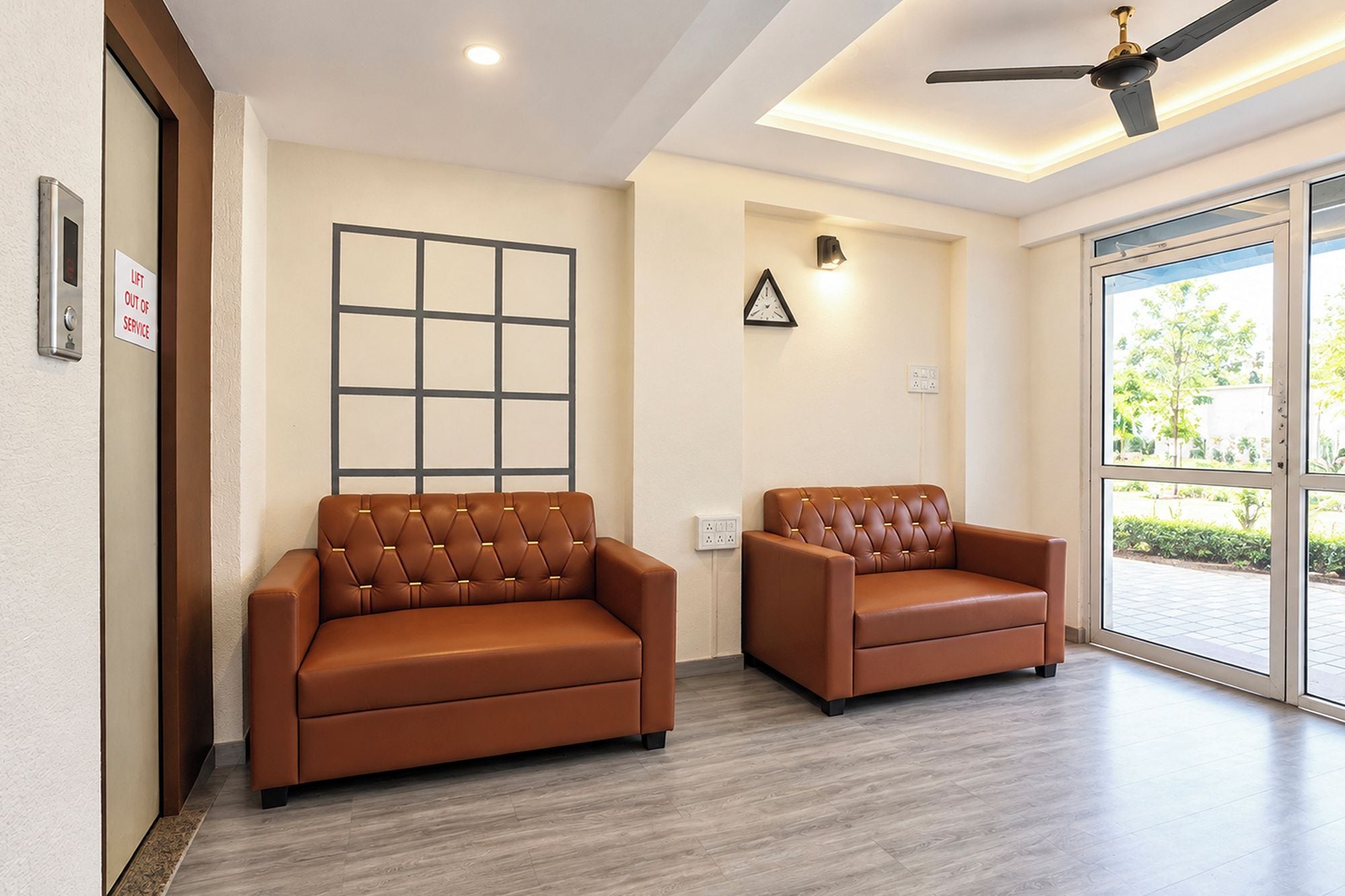 A waiting area with two leather sofas, a ceiling fan, a wall-mounted clock, and a large window with a view of greenery.