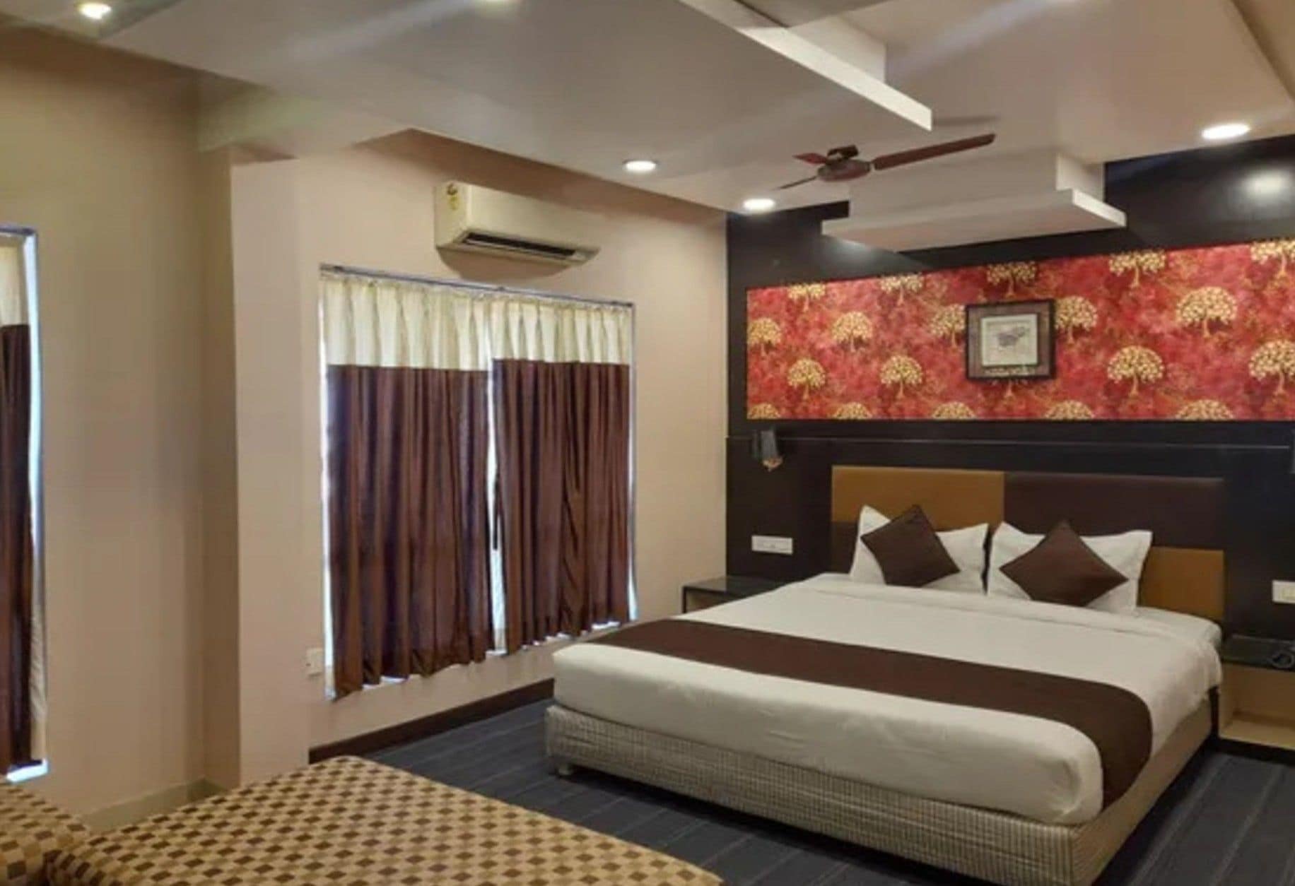 Una habitación de hotel con una cama grande, un ventilador de techo, un equipo de aire acondicionado, una ventana con cortinas y una pared con diseño.
