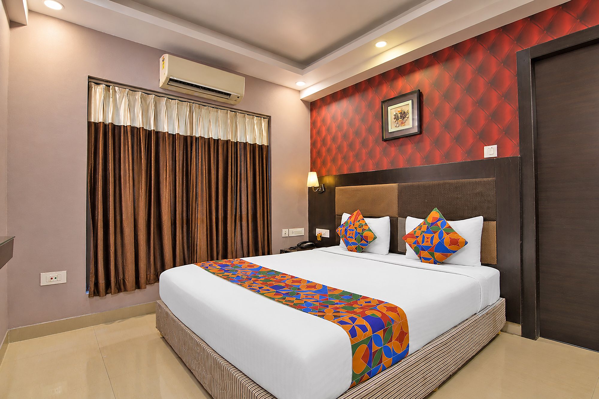 Una habitación de hotel con una cama grande, cabecera, aire acondicionado montado en la pared, ventana con cortinas y una pared con diseño geométrico.