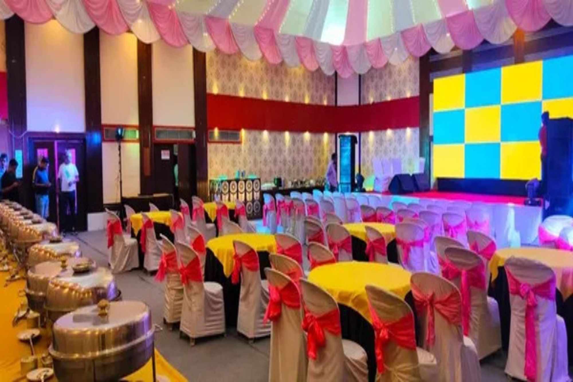 Une salle de banquet avec des tables rondes, des chaises ornées de rubans rouges et une scène dotée d’un décor coloré.