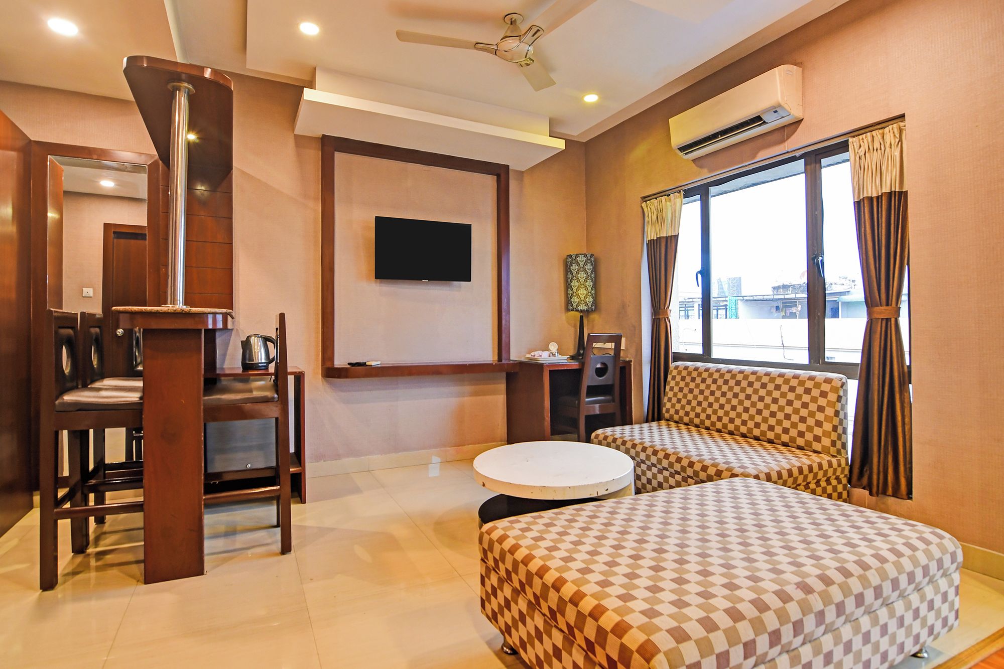 A hotel room with a checkered sofa, a dining table, a TV mounted on the wall, and an air conditioning unit.