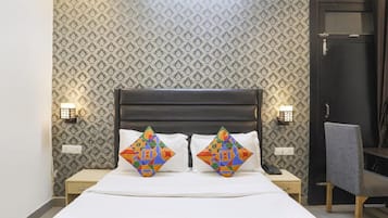Une chambre d’hôtel avec un papier peint à motifs, une tête de lit sombre, deux lampes de chevet, un bureau avec une chaise et un lit agrémenté d’oreillers géométriques aux couleurs vives.