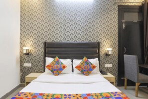 A hotel room with a patterned wallpaper, a dark headboard, two bedside lamps, a desk with a chair, and a bed with colorful geometric pillows. - FabHotel The Diamond (Noida)