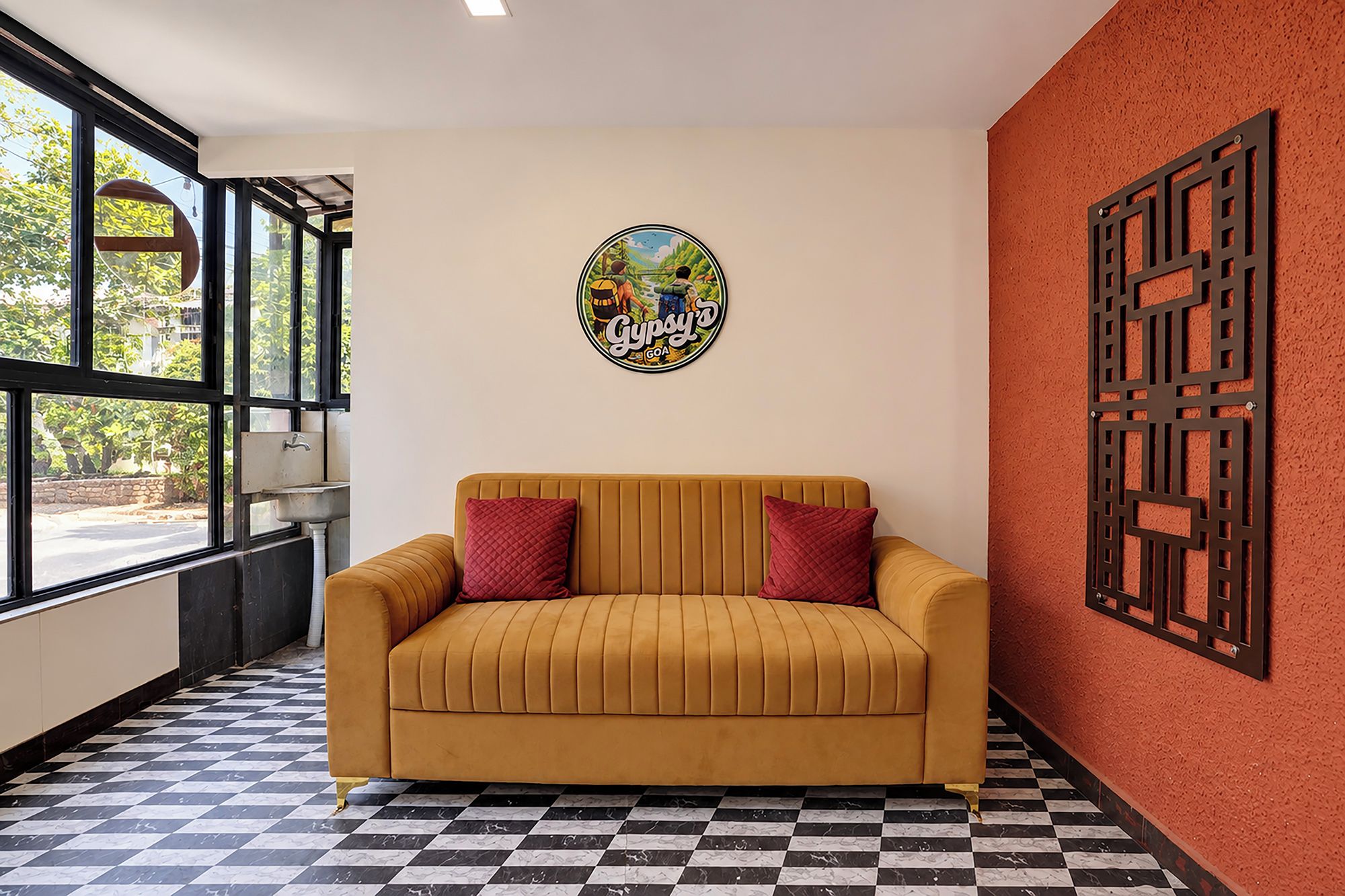 Una habitación con piso a cuadros, un sofá con cojines rojos, una pared con un letrero decorativo y una ventana con vista a follaje.