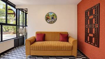 Una habitación con piso a cuadros, un sofá con cojines rojos, una pared con un letrero decorativo y una ventana con vista a follaje.