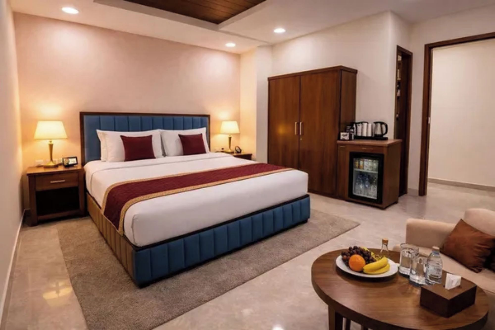 A hotel room with a large bed, bedside lamps, a wardrobe, a minibar, and a small table with fruits and a glass.