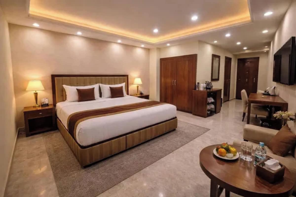 A hotel room with a large bed, bedside lamps, a desk, a chair, and a dining table with a plate of fruit.