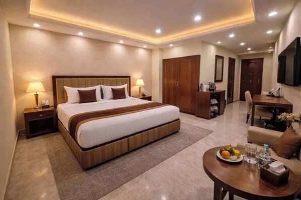 A hotel room with a large bed, bedside lamps, a desk, a chair, and a dining table with a plate of fruit.