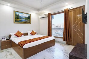Habitación de hotel con cama, mesita de noche, un cuadro, ventana con cortinas y vista a edificios.