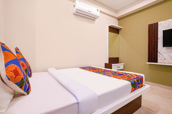A hotel room with a bed, a bedside table, an air conditioning unit, a mirror, and a television mounted on the wall.