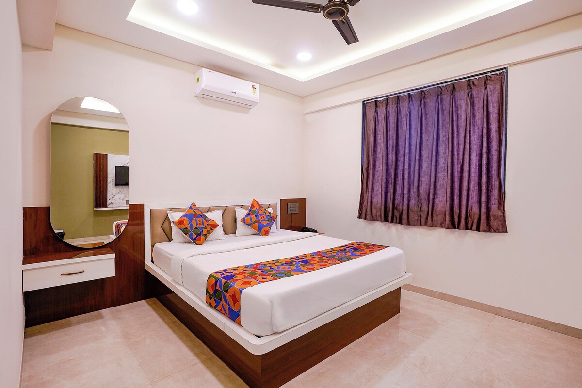 Habitación de hotel con cama, mesita de noche, espejo, unidad de aire acondicionado y ventana con cortinas.