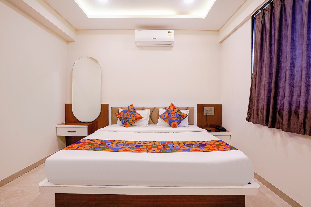 A hotel room with a bed, a bedside table, a nightstand, a mirror, a window with curtains, and an air conditioning unit.