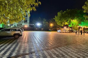 Un espacio pavimentado con vehículos y personas, un edificio con ventanas iluminadas y árboles de hojas verdes.