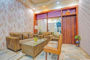 A living room with a sofa set, a glass coffee table, and a chair. There is a wooden door with a decorative panel and a small plant on the floor. - FabHotel Amantran (Kanpur)