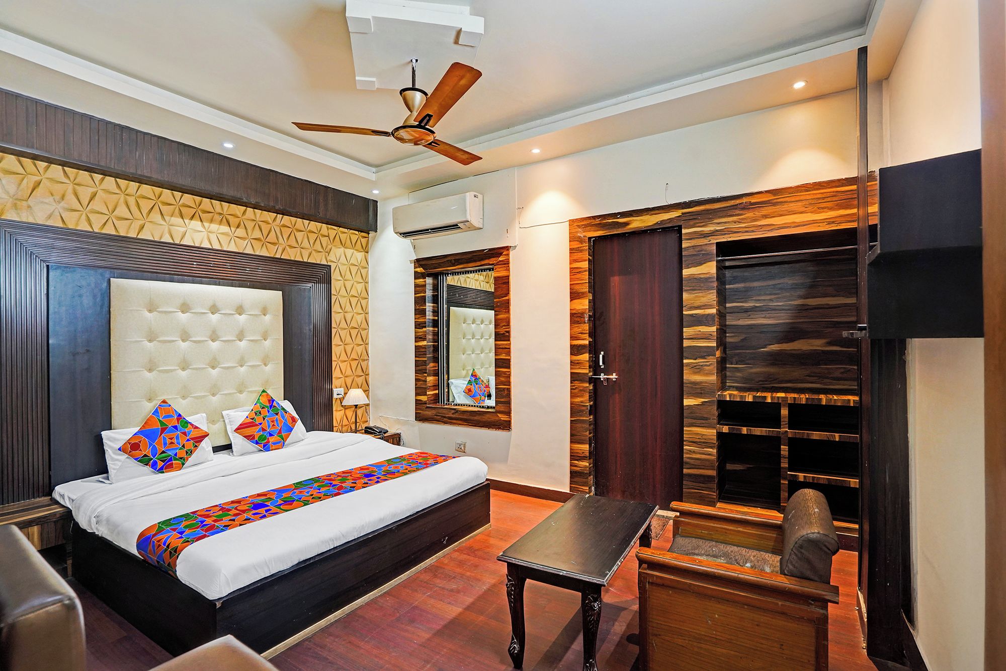 A hotel room with a large bed, a ceiling fan, a wooden door, and a built-in TV shelf.