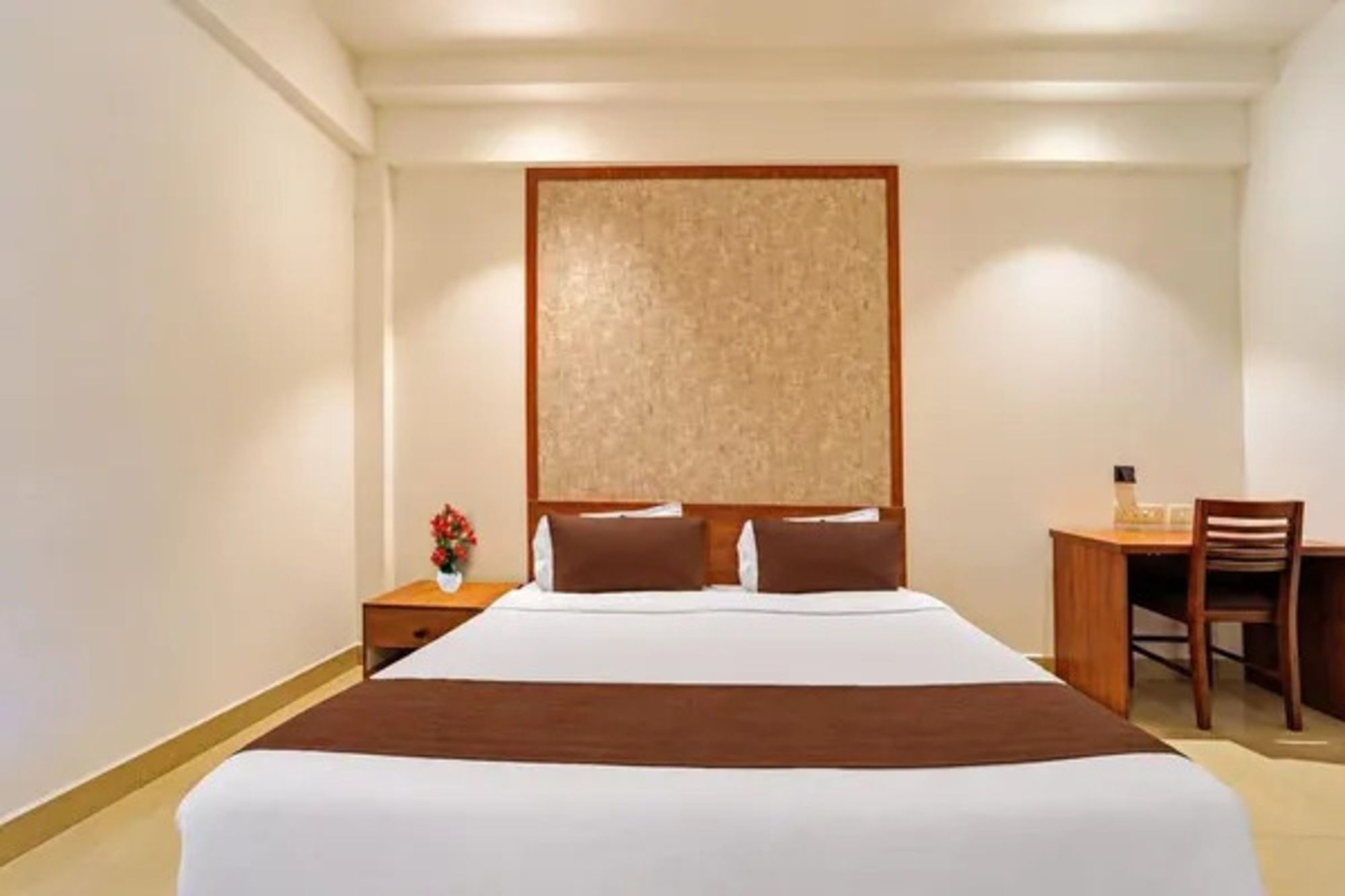 A hotel room with a large bed, a wooden headboard, a nightstand with a vase of flowers, and a desk with a chair.