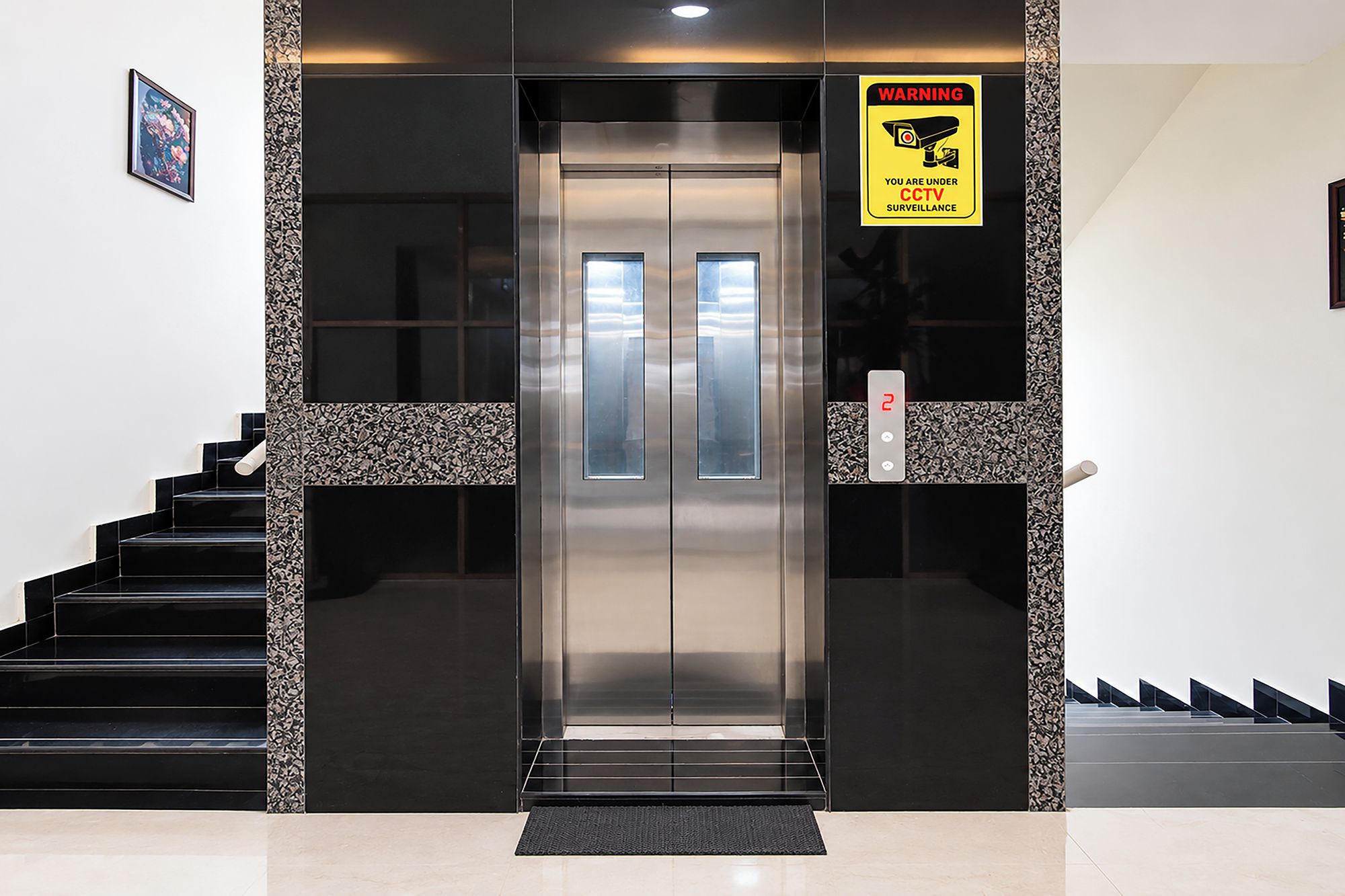 Un ascensor con un letrero de advertencia de circuito cerrado de televisión, una escalera con escalones negros y una pared con diseño.