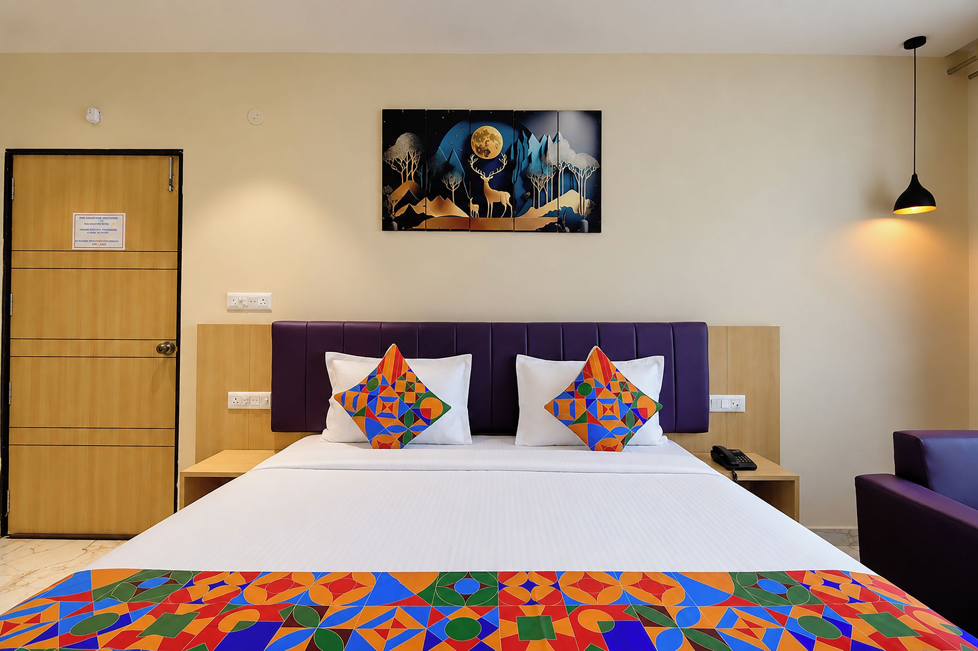 Quarto de hotel com cama, duas almofadas, mesa de cabeceira, telefone, abajur e uma obra de arte na parede.