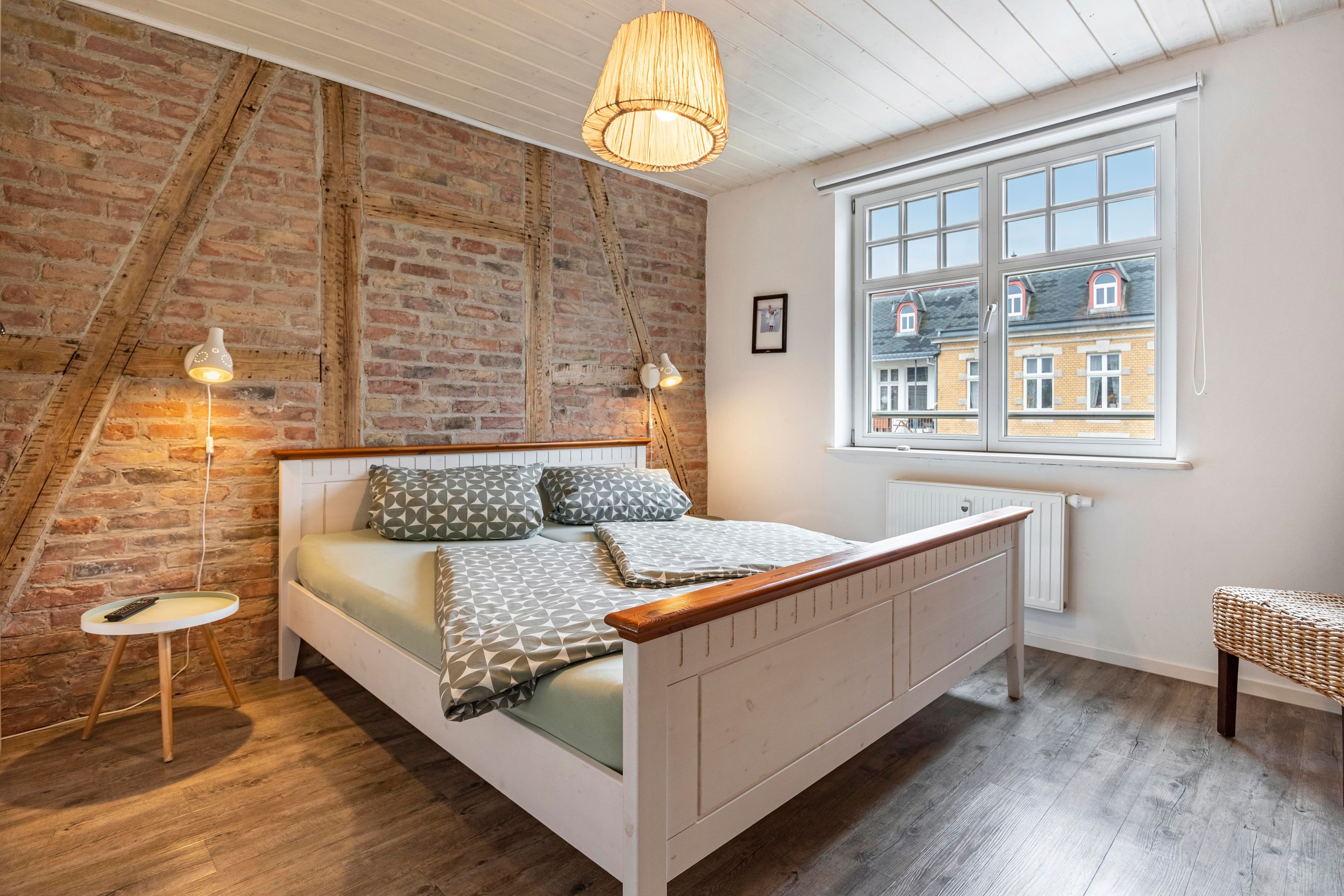 Ein Schlafzimmer mit schrägem Dach, freiliegenden Backsteinwänden, einem großen Fenster, einem Bett mit gemusterten Betttextilien und einem kleinen Nachttisch.