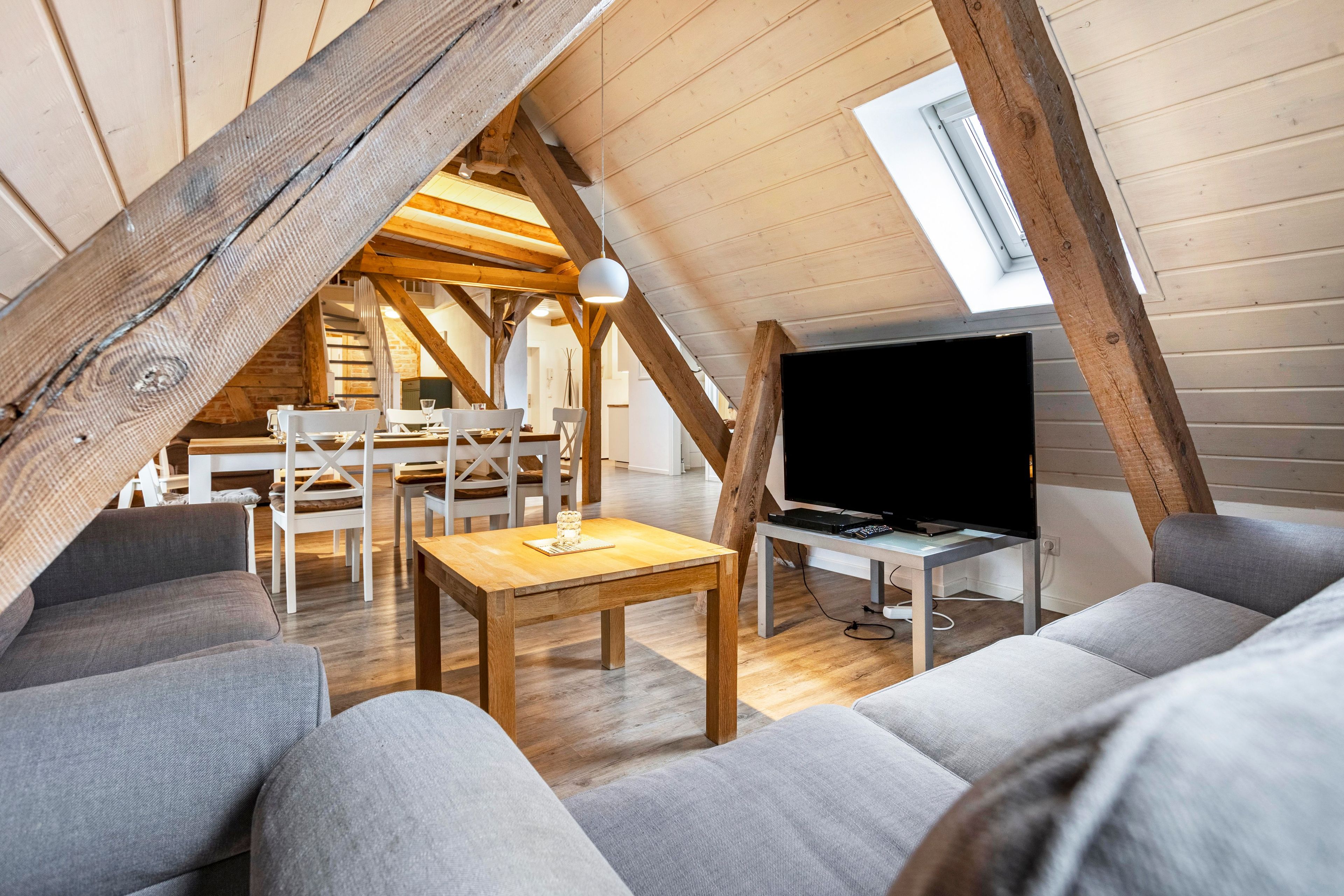 A loft-style room with a wooden ceiling, a dining area with white chairs, a television on a stand, and grey sofas.