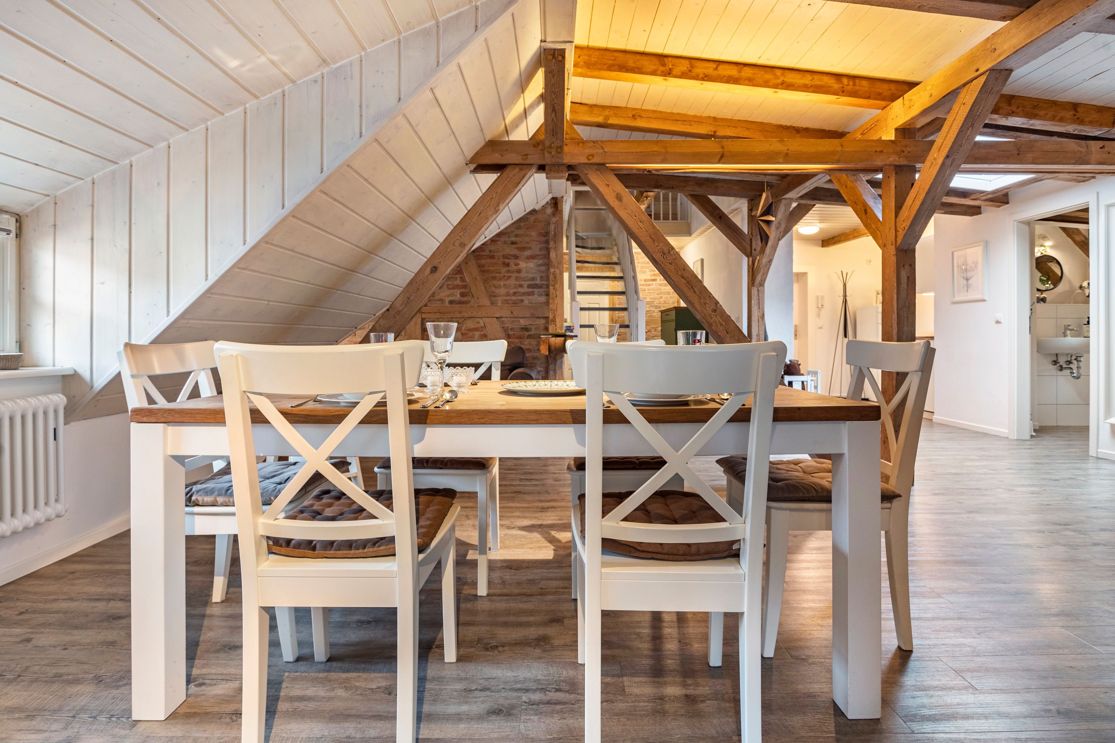 Un coin repas avec une table en bois et des chaises blanches, un escalier et une vue sur une salle de bain.