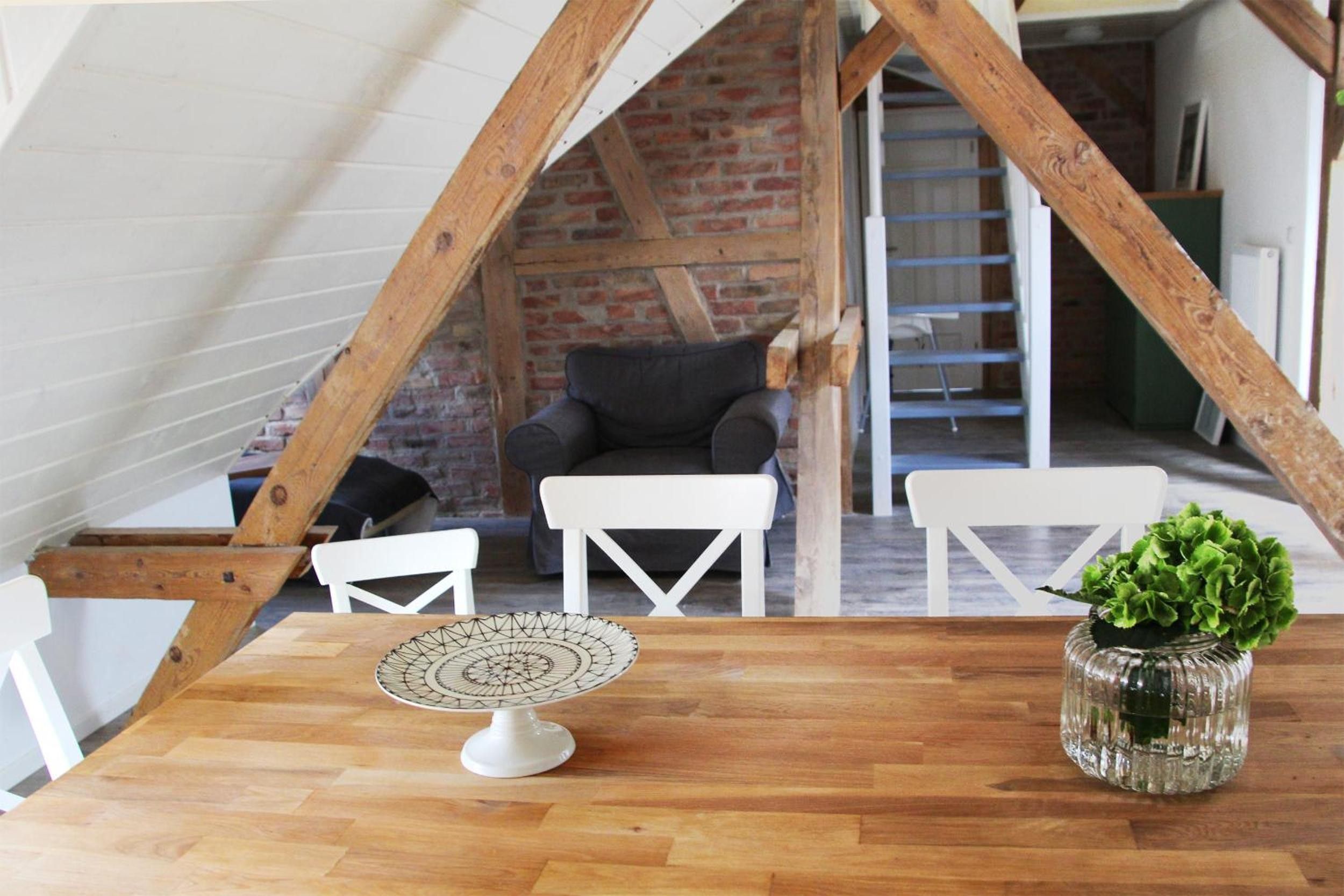 Une pièce de style « loft » avec des poutres en bois apparentes, un mur en briques et un plancher en bois. On y trouve une table blanche ornée d’une assiette décorative, une chaise blanche et une petite plante en pot.